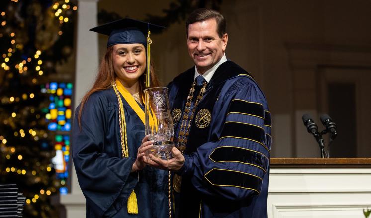 A man and a women in graduation regalia holding a clear award in a chapel