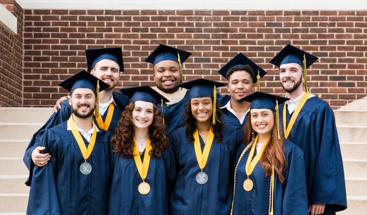 A group of graduates in navy cap and gowns smiling while standing on steps in front of a brick building.
