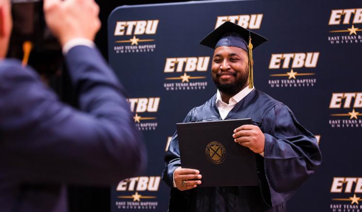 A man smiling at a camera in his graduation regalia holding his diploma up with a navy backdrop