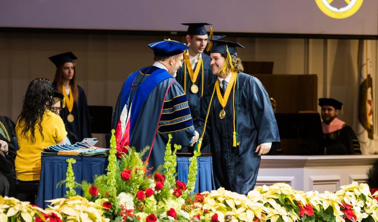 Two men shaking hands smiling in their graduation regalia on a stage with other people in the background