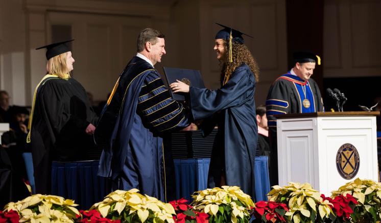 A man and woman in graduation regalia shaking hands handing off a diploma on stage with a man and woman in the background