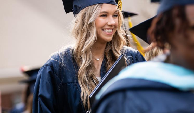 Women smiling in graduation regalia outside with another woman blurred in the foreground