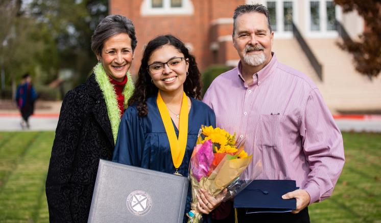 Family of two women and one male with the women in the middle in her graduation regalia holding her diploma and flowers outside