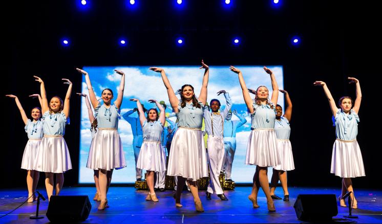 A group of dancers in light blue tops and white skirts performing on stage with arms raised, against a backdrop of a sky and clouds.