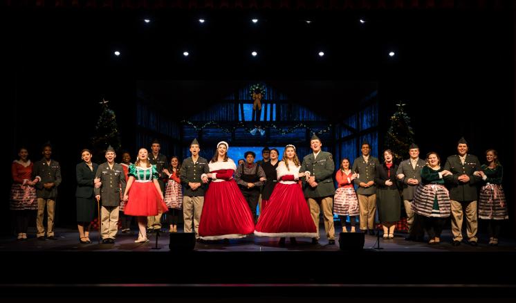 A group of people on a stage, wearing formal and traditional attire, lit by spotlights in a dark theater.