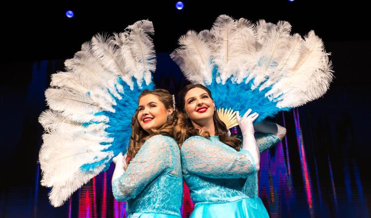 Two performers with large feathered fans on stage, wearing matching blue dresses.