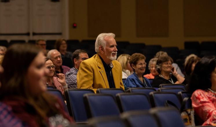 An audience sits in blue chairs in an auditorium, with a white-haired man in a yellow jacket standing among them.