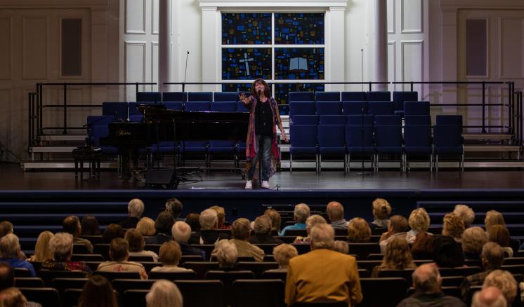  Person speaking on a stage in front of a seated audience in an auditorium.