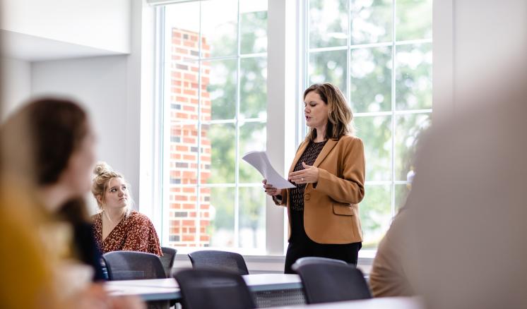  Woman standing and presenting in a classroom with seated attendees and large windows in the background.