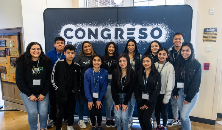 Group of students standing in front of event banner smiling at the camera 