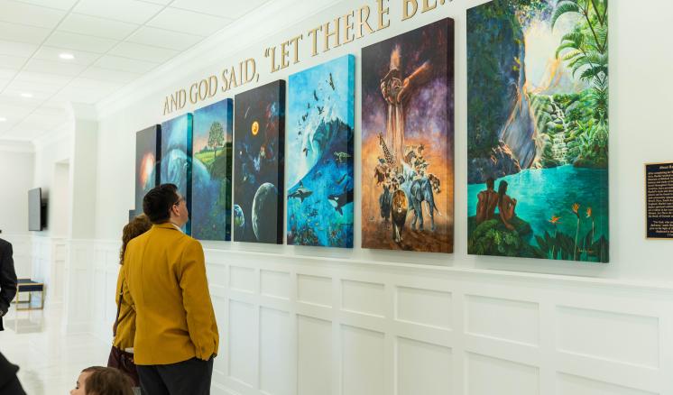 People viewing a row of colorful paintings in a bright gallery space with gold lettering above.