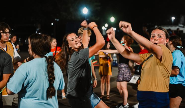 People dancing energetically at a nighttime outdoor event, illuminated by streetlights.