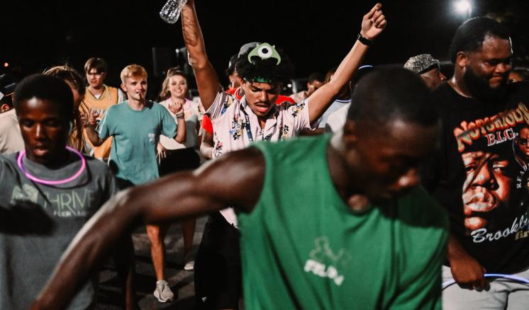 A group of people dancing and celebrating outdoors at night, with one person in a green shirt prominent in the foreground.