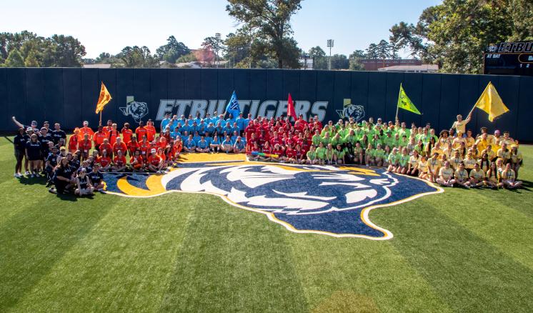 Groups of people in colored uniforms forming a semi-circle on a field with an "ETBU TIGERS" logo in the background.