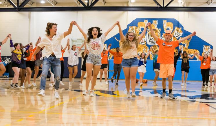 A group of people in a gymnasium holding hands and raising their arms in celebration, with a large mural in the background.