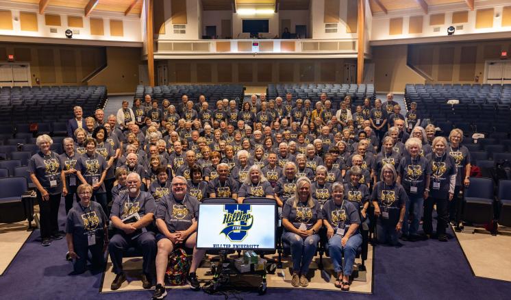 Hilltop U 2023 Large group of people in an auditorium wearing matching grey t-shirts, with a screen in front displaying a logo.