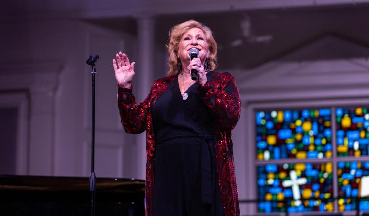 A woman wearing a red jacket singing on stage with hand outstretched.