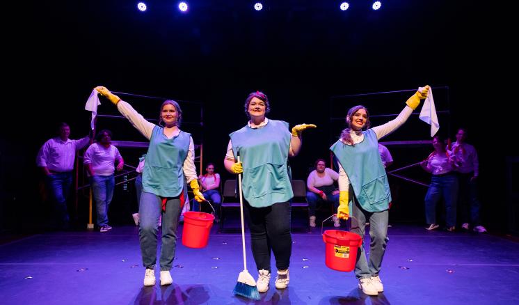 Three women standing in foreground Working:The Musical theatre production.