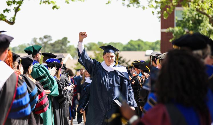 A graduate in a navy blue gown and cap raises a hand while walking through an aisle lined with people in similar attire during a graduation ceremony.