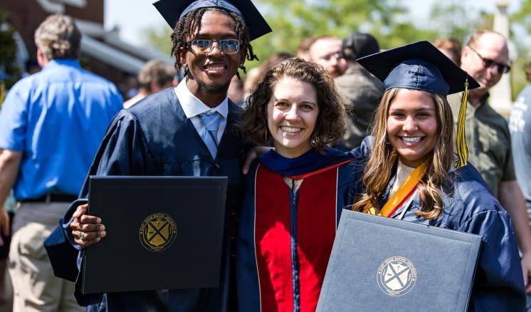 Three people at a graduation ceremony, two in caps and gowns holding diplomas, and one in an academic robe.