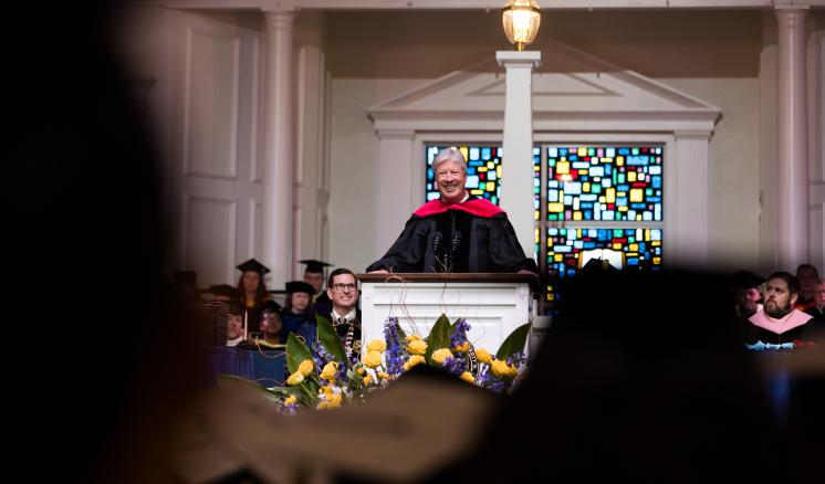 A man in academic regalia speaking behind podium on a stage with a crowd in the foreground