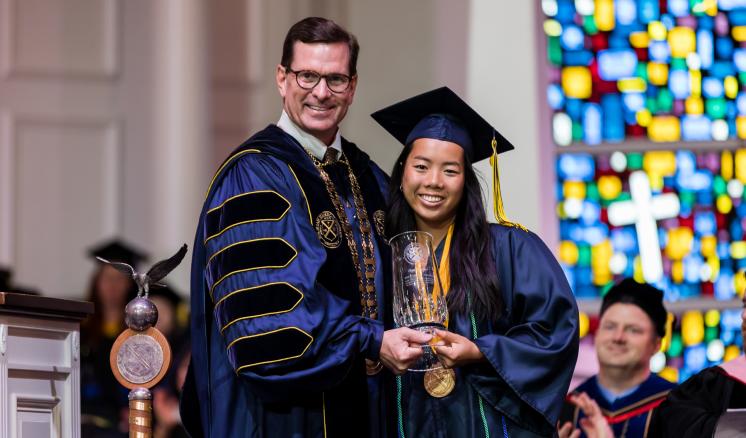 A graduate and a faculty member at a graduation ceremony, both smiling and holding an award.