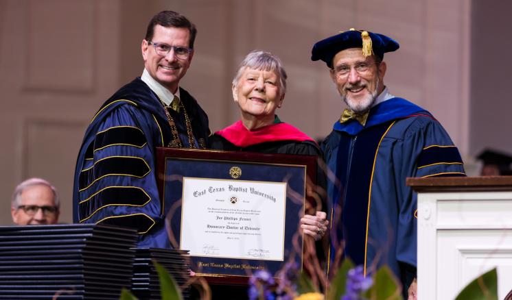 Three people in academic regalia holding a diploma, smiling.