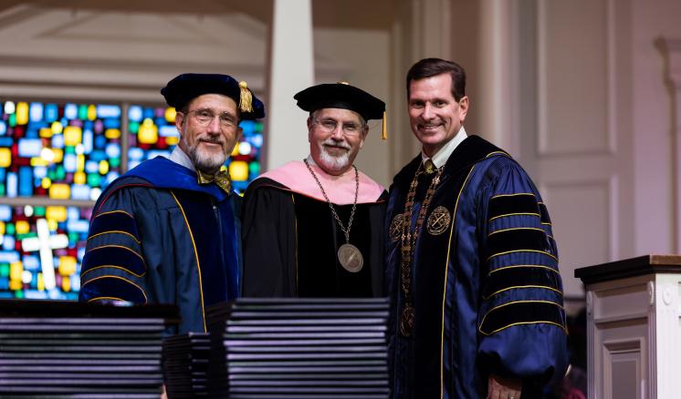 Three men in academic regalia standing and smiling; stained glass window in the background.