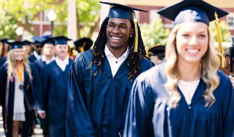 A group of graduates in navy blue gowns and caps walking outdoors.