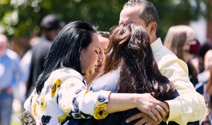 A group of four people hugging outdoors with a blurred background of trees and people.