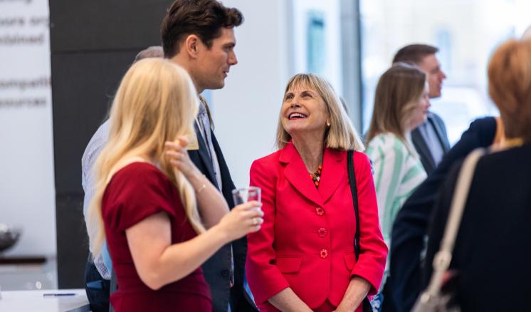 People at a social or professional gathering, with a woman in a maroon dress, man in a grey suit, and woman in a red blazer in the foreground.