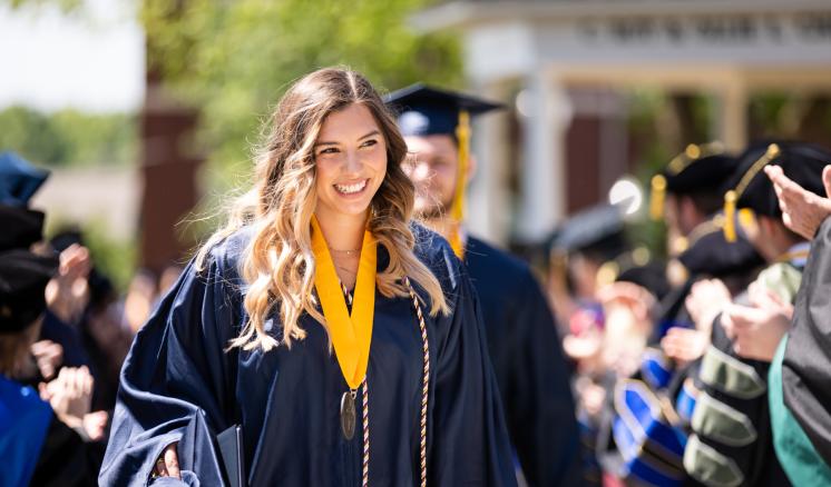 A young woman in graduation attire smiles as she walks during a graduation ceremony.