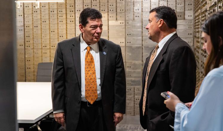 Two men in suits talking in front of safety deposit boxes inside a bank vault. A third person with a smartphone is partially visible on the right.