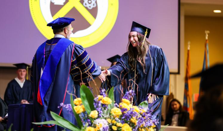 A graduate and a faculty member shake hands onstage at a graduation ceremony, with flowers in the foreground and a projected emblem in the background.