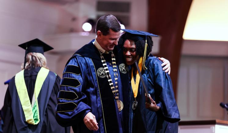 Two people in academic regalia share a congratulatory moment at a graduation ceremony.