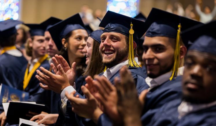 Graduates in navy caps and gowns clapping during a ceremony.