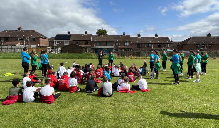 img_1 Children seated on a grassy field surrounded by adults in colorful shirts, with houses and a partly cloudy sky in the background.