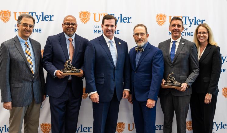 Group of five males and one female smiling at camera in front of a white UT Tyler backdrop