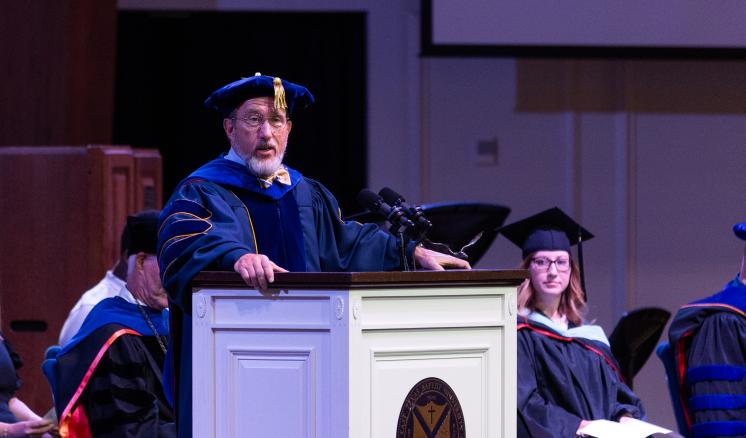 A man in graduation attire speaking behind a podium inside with people sitting in the background