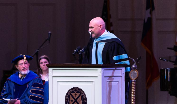 A man in graduation attire speaking behind a podium inside with people sitting in the background