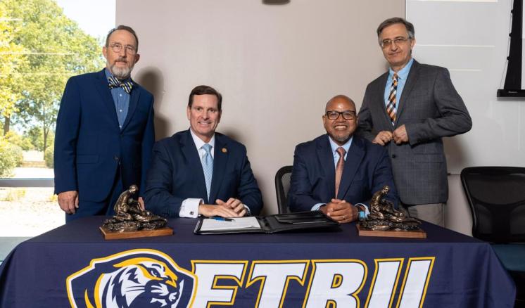 Four males inside smiling at camera behind table with navy blue ETBU tablecloth over it with awards and a journal on the table