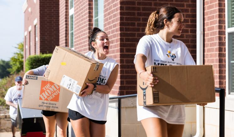 A line of people holding moving boxes entering brick building