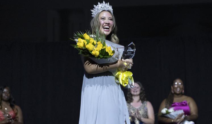 A female inside smiling in a ballgown with a crown on and flowers and an award in her hands