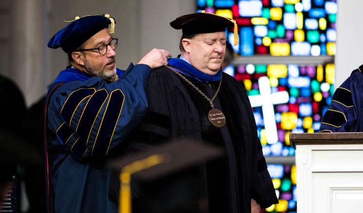 Man in academic regalia putting a medal around another mans neck in academic regalia