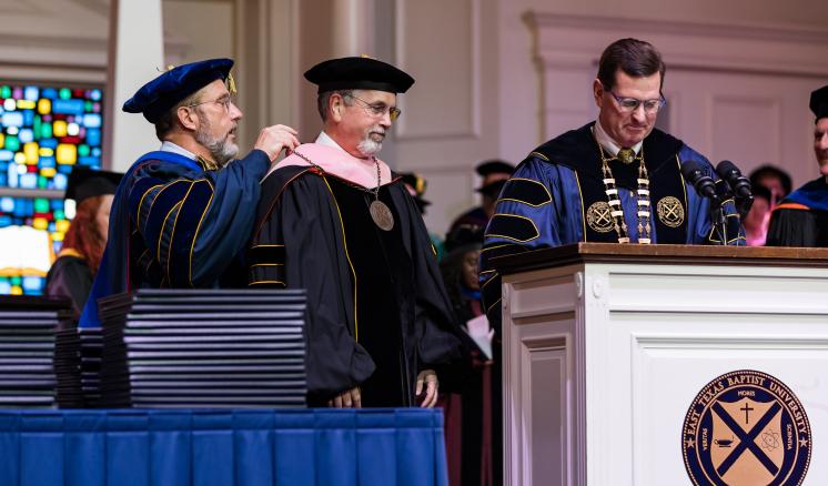 Man in academic regalia putting a medal around another mans neck in academic regalia with another man in academic regalia behind a podium