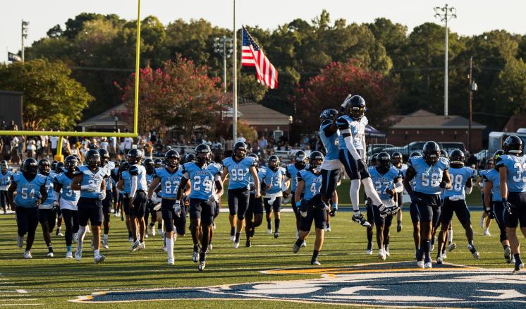 A group of football players in blue uniforms running down a football field toward the camera.