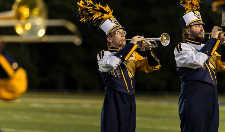 Two trumpet players in marching band uniforms playing music on a football field at night.