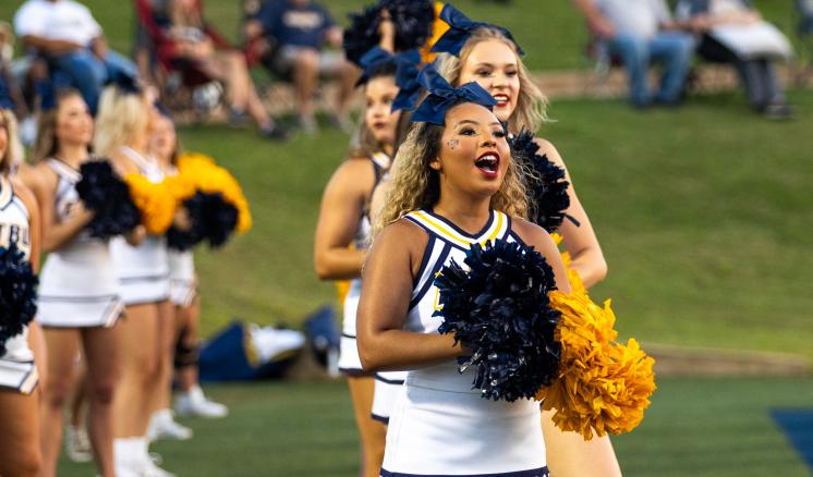 Cheerleaders yelling on the sidelines of a football game with gold and navy pompoms.