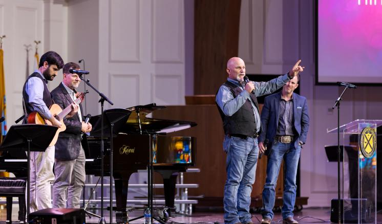 A man talking on stage with arm extended with males in the background