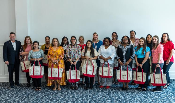 Group of females and one man smiling at the camera holding white and pink tote bags inside